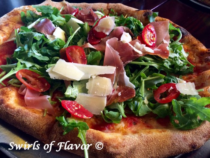 Grilled Prosciutto Parmesan & Arugula Pizza is topped with a bed of baby arugula greens lightly tossed in a white balsamic vinaigrette dressing. Baby arugula, tomatoes and prosciutto combine to make each bite the perfect combination of summer flavors.