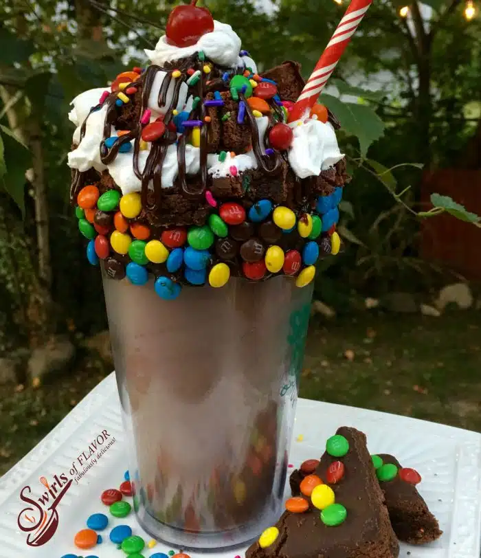 The Ulitmate Brownie Milkshake is filled with chocolate ice cream and brownie bits with a fudgy rim of mini m&m's and brownie chunks topped with creamy whipped cream, mini marshmallows, rainbow sprinkles, a drizzle of hot fudge sauce and a cherry on top! milkshake | over the top | chocolate | chocolate milkshake | over the top milkshake | brownie | M&M