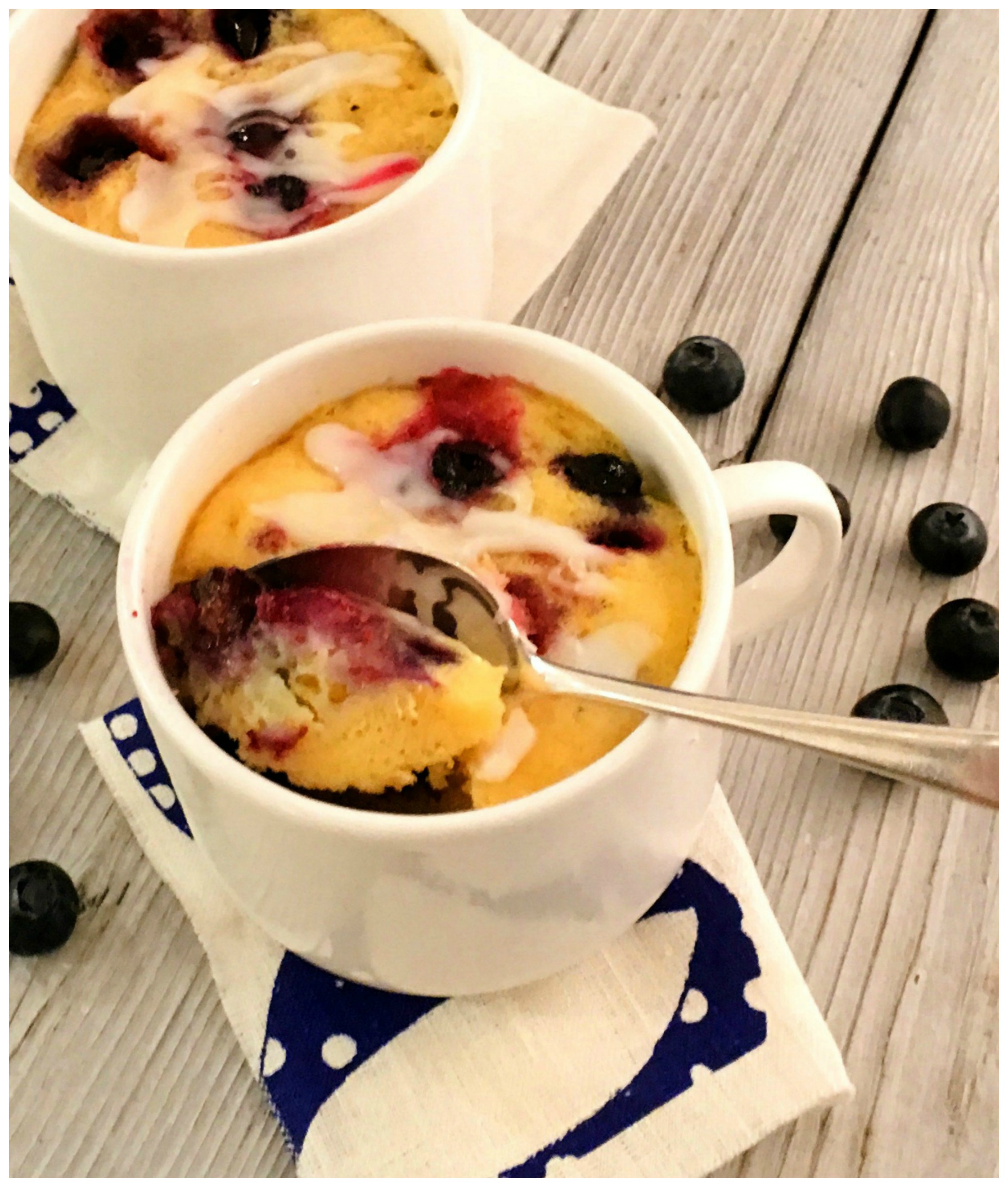 two blueberry mug cakes with spoon and fresh blueberries