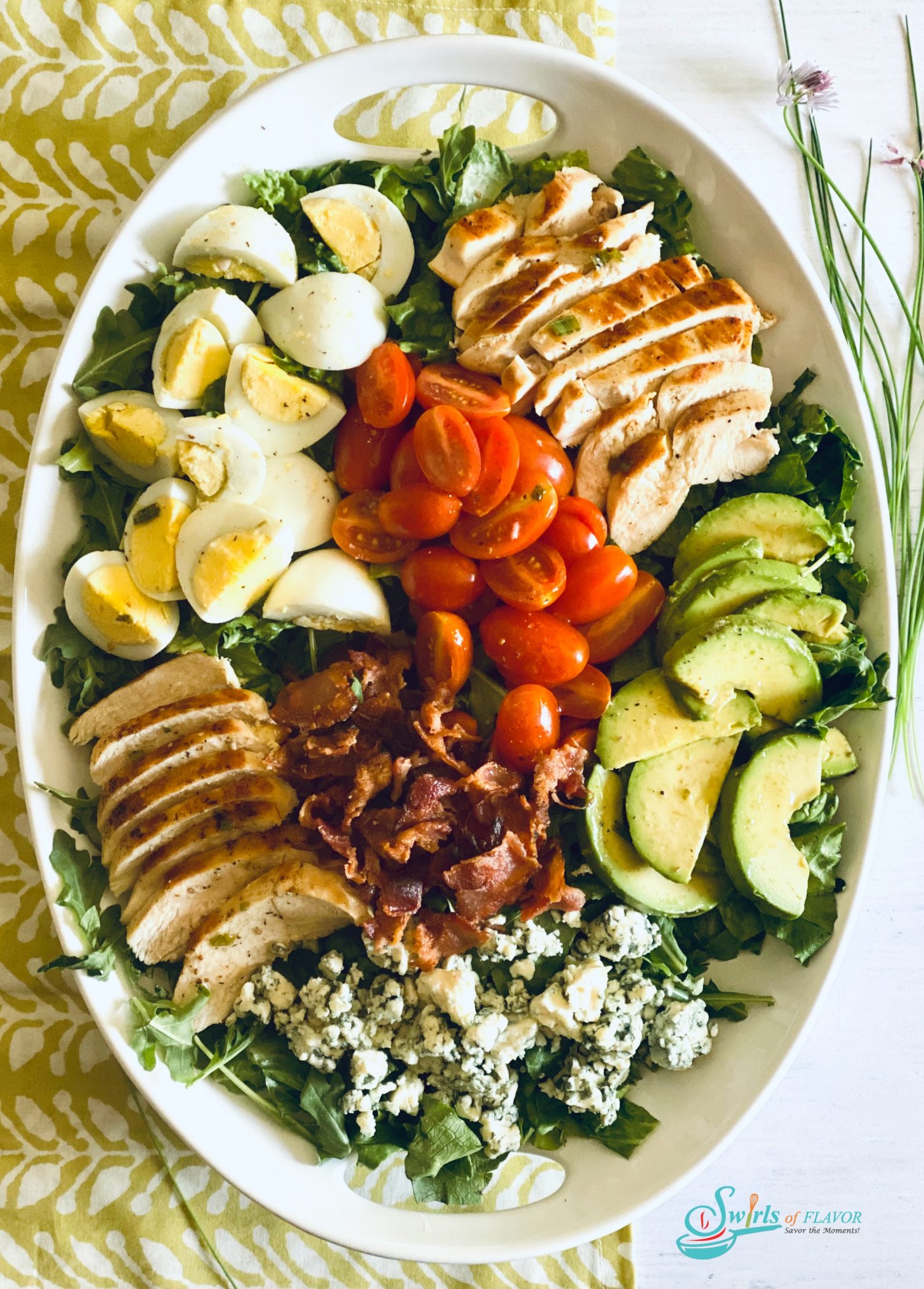 Overhead photo of a Cobb Salad in white oval platter on a green and white napkin with fresh chives