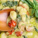 close up of pesto cream sauce with gnocchi and tomatoes and wooden spoon and text overlay