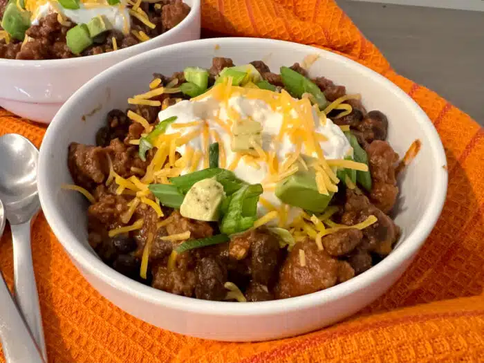 a bowl of beef chili with sour cream, shredded cheese and sliced green onions toppings