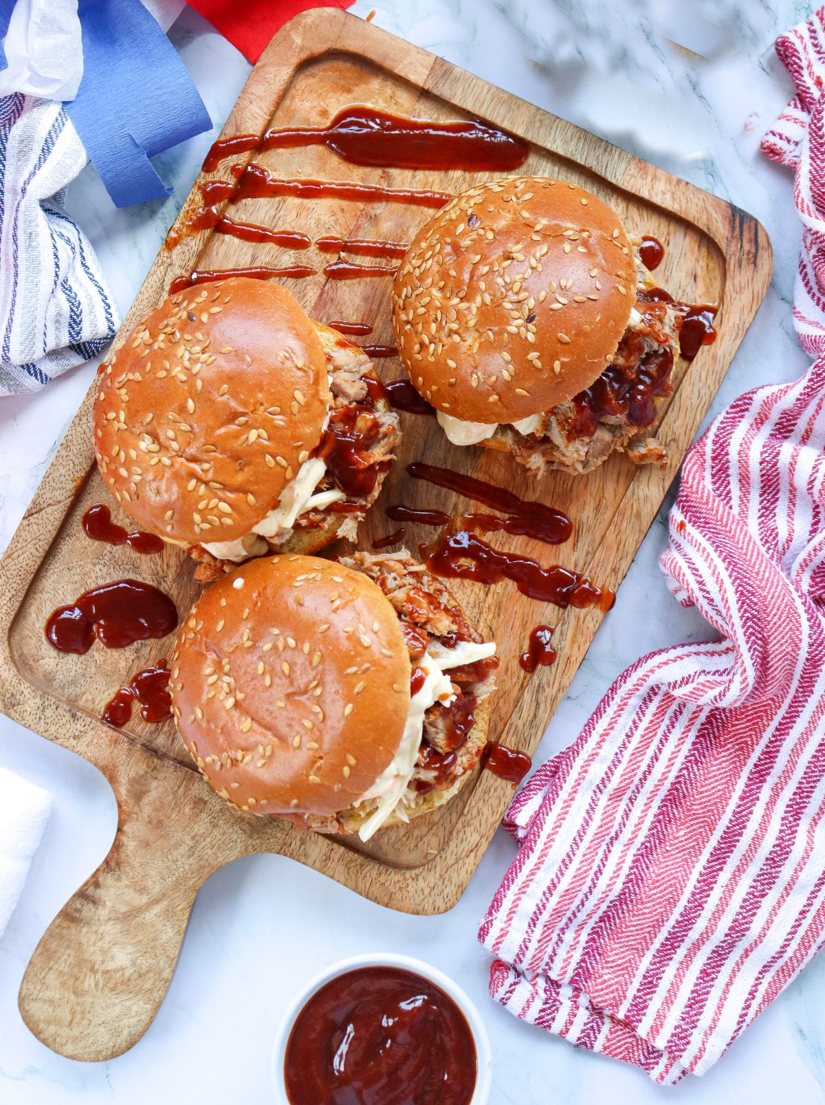 overhead photo of three pulled pork sandwiches on a wooden board with colorful napkin
